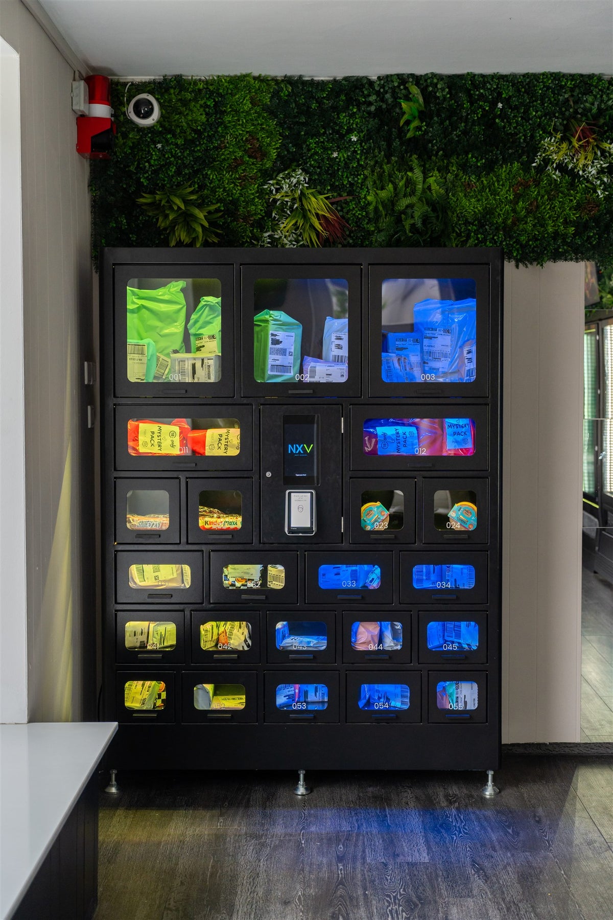 Schwarzer Fächerautomat mit Secret Packs in unterschiedlich großen, beleuchteten Fächern vor einer Pflanzenwand.