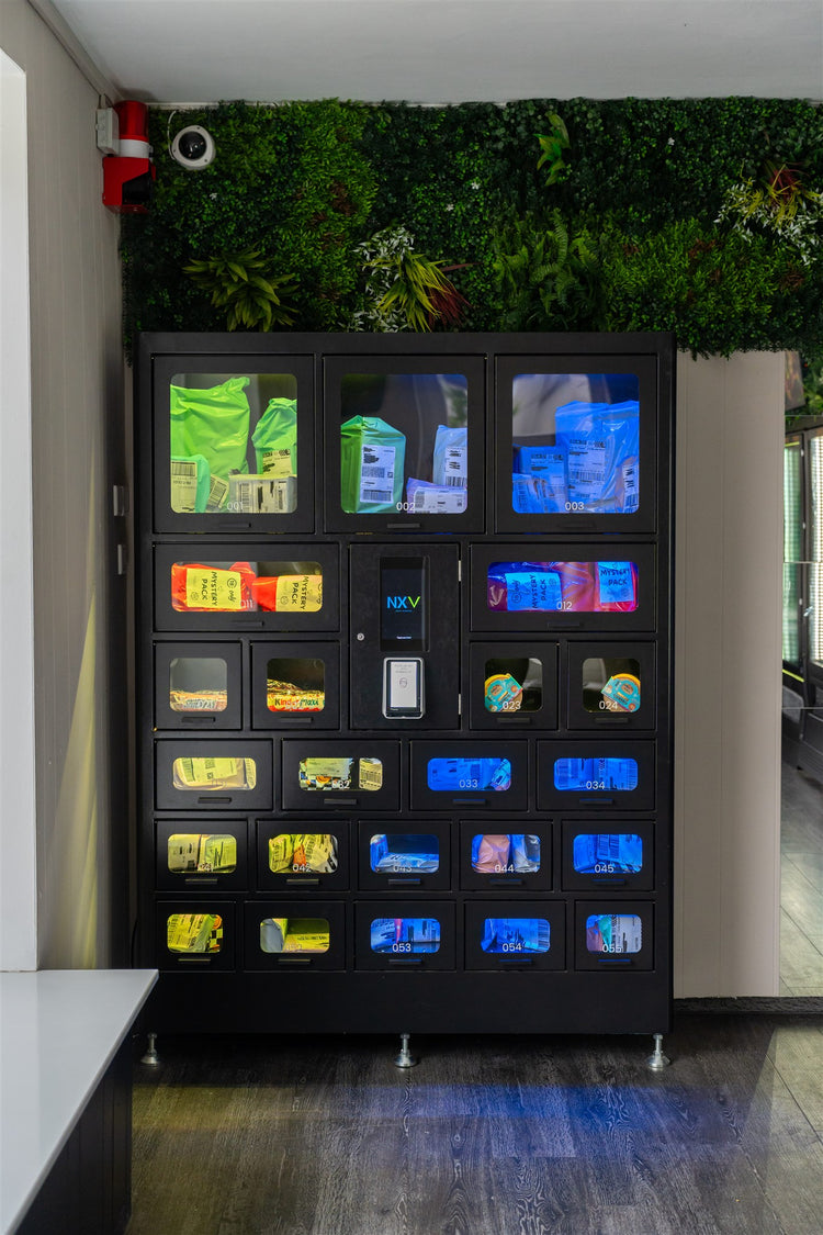 Schwarzer Fächerautomat mit Secret Packs in unterschiedlich großen, beleuchteten Fächern vor einer Pflanzenwand.